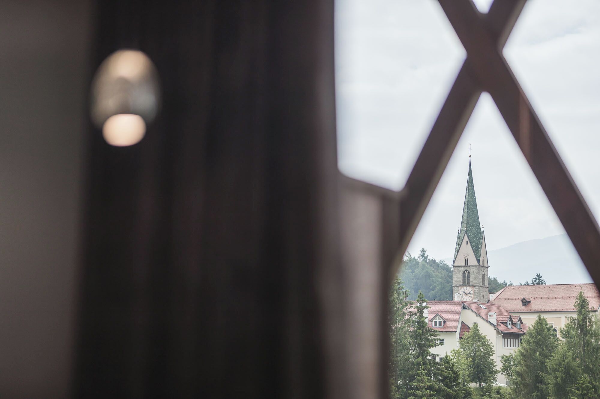 Une vue à travers une fenêtre ornée d’un cadre géométrique, mettant en évidence une église dotée d’un flèche haute et d’un clocher.
