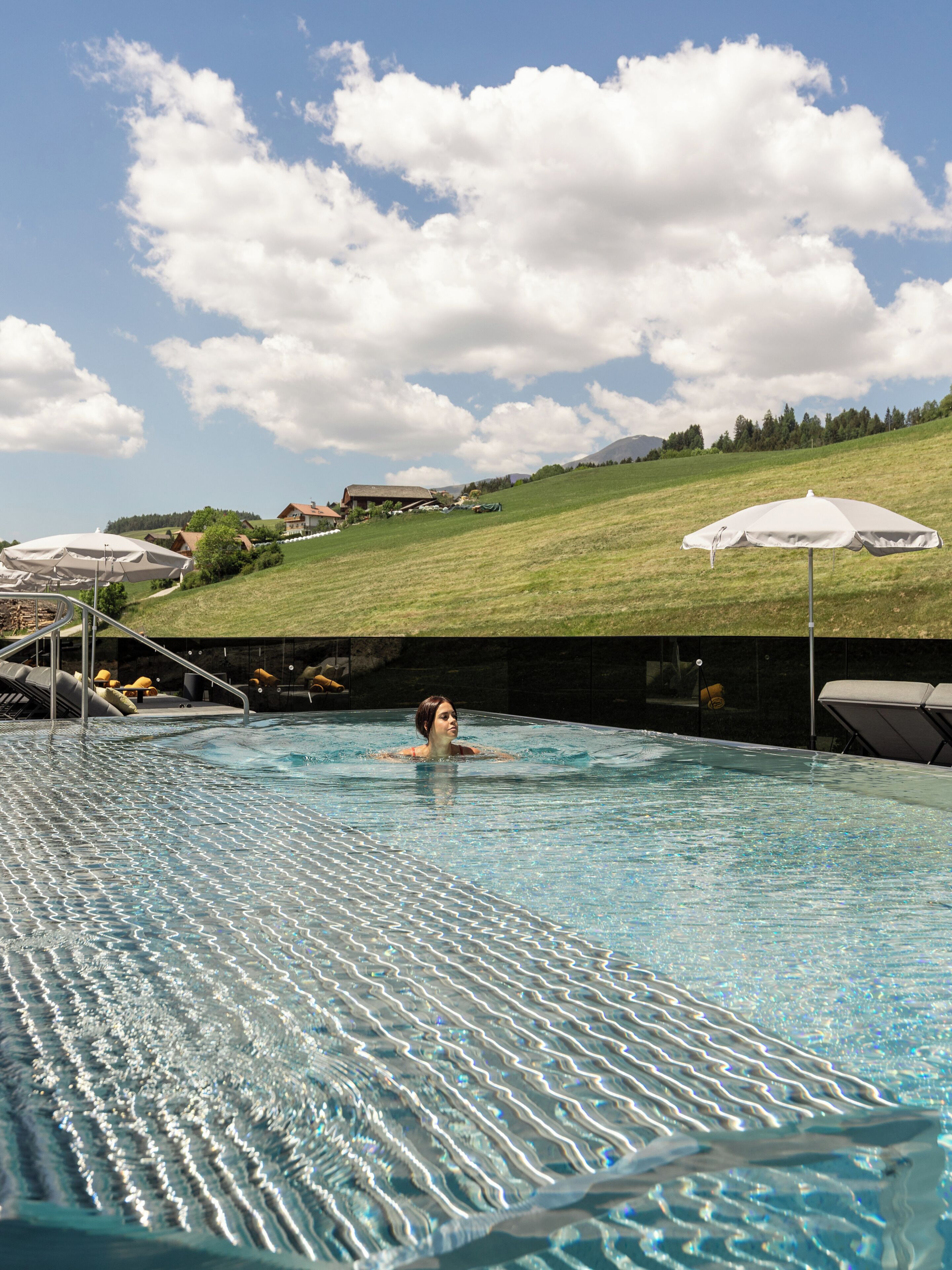Una piscina con una persona nadando, rodeada de sillas de descanso y sombrillas, y un paisaje montañoso al fondo.