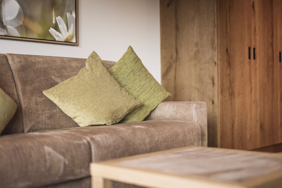 Een bruine leren bank met twee groene kussens en een houten salontafel in een woonkamer met een houten garderobe en een lijst met een afbeelding erop.