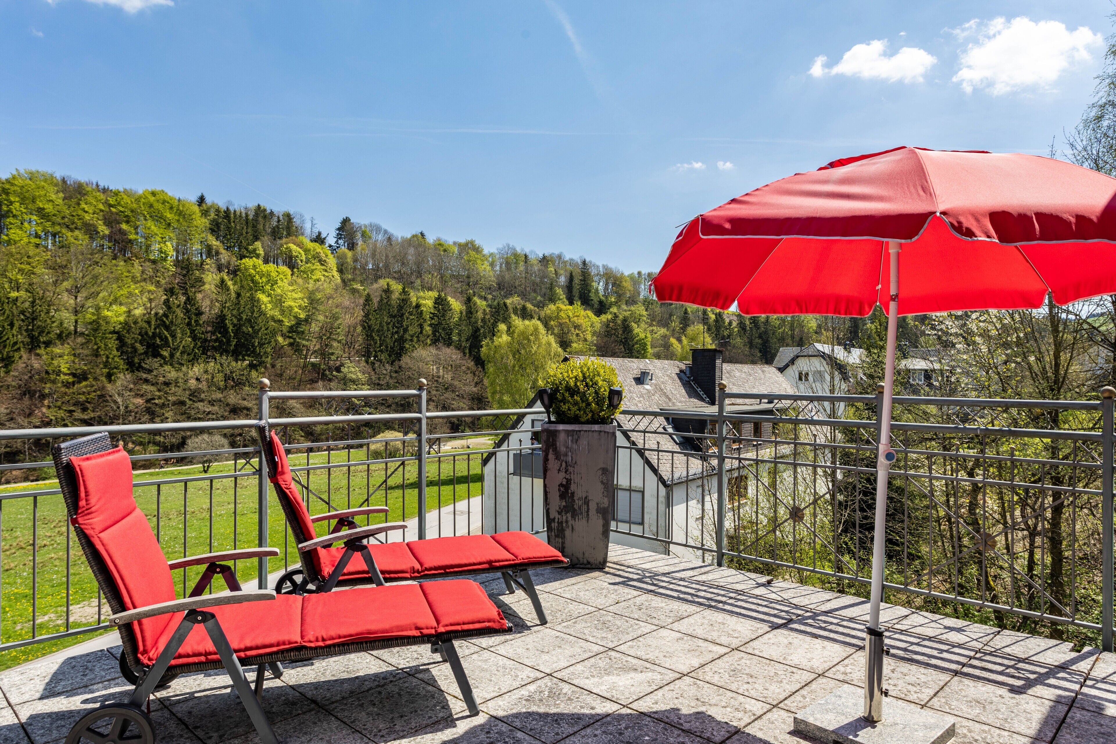 A balcony with two red lounge chairs and a red umbrella, overlooking a green landscape with trees and a building in the background.
