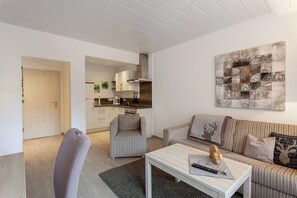 A modern living room with a sofa, armchair, and coffee table. A kitchen is visible in the background.