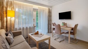 Quarto de hotel moderno com sofá, uma mesinha, TV fixada na parede e um espaço de refeições com cadeiras.