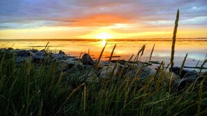 Een zonsondergang boven kalm water, met rotsen en gras op de voorgrond.