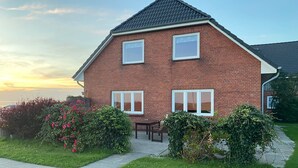 Een herenhuis met twee verdiepingen, gebouwd uit baksteen, met een geplaveid opritje, een klein terras en uitzicht op zee bij zonsondergang.