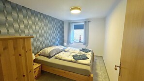 A bedroom with a bed, a wooden nightstand, a window with curtains, and a wall with a textured pattern. - Apartment 'Ferienwohnung Uthlande' with Shared Terrace, Shared Garden and Wi-Fi (Langeneß)