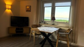 Quarto com televisão, mesa com toalha de lugar, duas cadeiras, janela com vista e luminária.