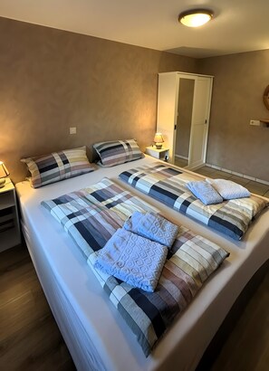 Uma cama de casal com colcha listrada e almofadas, mesa de cabeceira com abajur, guarda-roupa e luminária de teto.