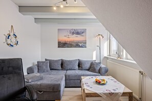 Un salon moderne avec un canapé d’angle, une table à manger ornée de fruits, un luminaire fixé au mur et un tableau représentant un coucher de soleil.