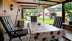 Un espacio de estar al aire libre cubierto con una mesa y sillas, una lámpara colgante y vistas a un jardín y casas al fondo.