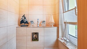 A bathroom with tiled walls, a window with blinds, and a shelf with decorative items including a sailboat model and framed pictures. - Apartment 'Familie Perlik' with Wi-Fi (Röbel)