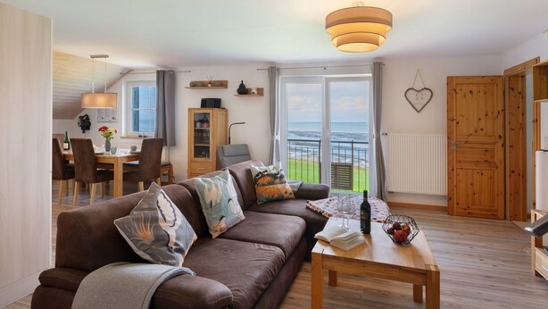 Ein Wohnzimmer mit einer Couch, einem Tisch mit Flasche und Gläsern, einem Essbereich mit Stühlen und einem Tisch sowie Meerblick durch große Fenster.