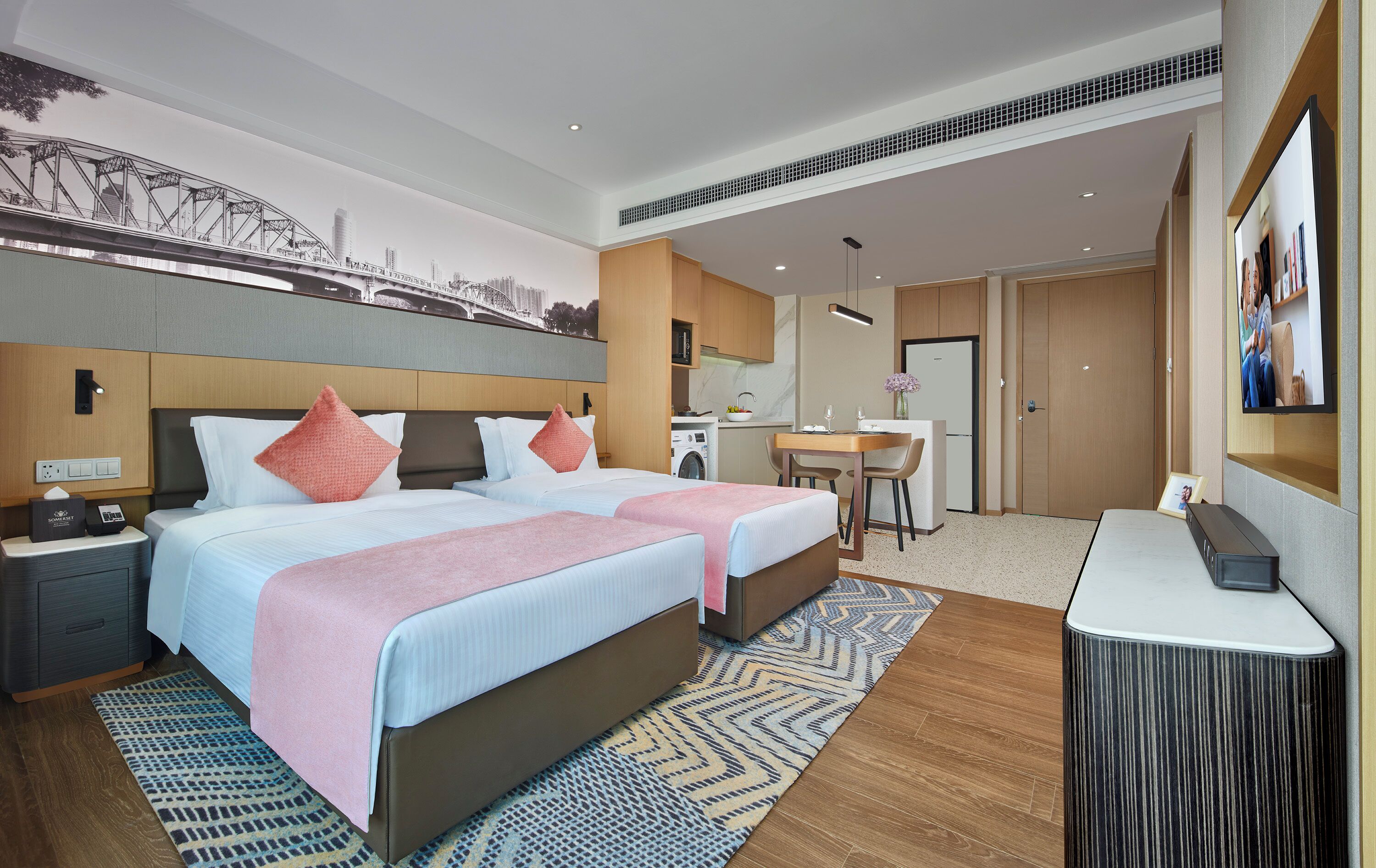 A hotel room with two beds, a desk, a small kitchenette, and a TV.