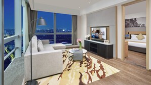 Ein modernes Hotelzimmer mit großem Fenster und Blick auf die Stadt, einer Couch, einem kleinen Tisch mit Blumenvase, einem Flachbildfernseher und einem Bett mit Nachttisch.
