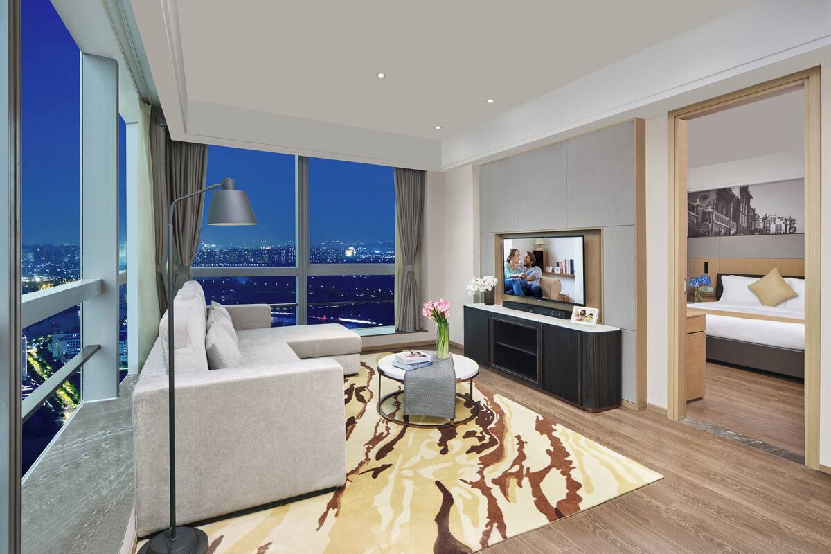 Ett modernt hotellrum med ett stort fönster som erbjuder utsikt över staden, en soffa, ett litet bord med en vas med blommor, en platt-TV och en säng med ett nattduksbord.