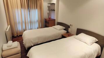 A hotel room with two beds, a small desk, a chair, and a window with curtains.