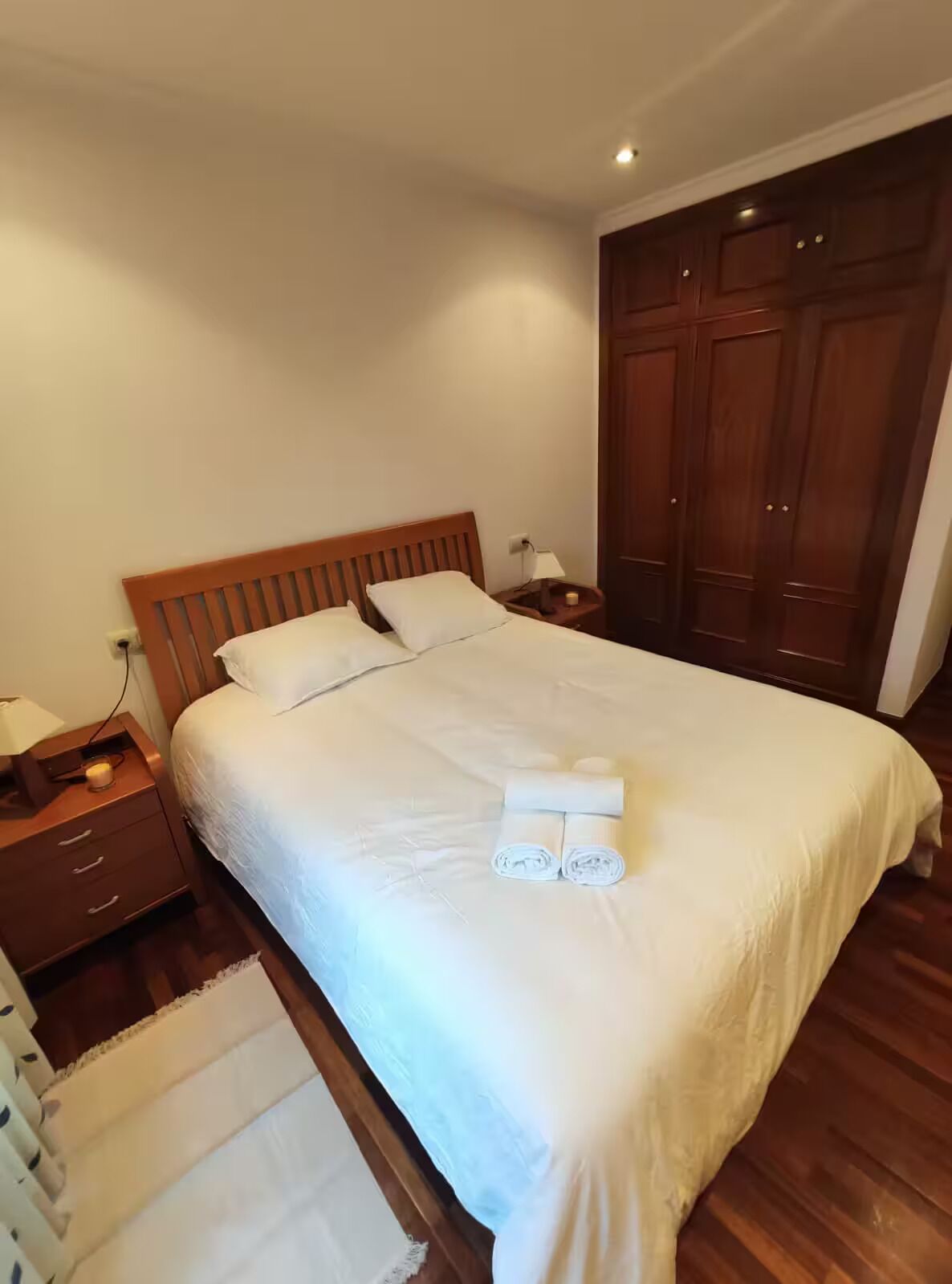 A neatly made bed with white linens, a wooden headboard, and a nightstand with a lamp and a candle.