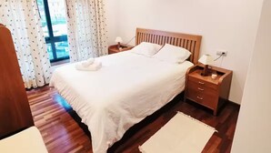 Une chambre à coucher bien aménagée, avec un grand lit, une tête de lit en bois, des tables de chevet et une vue sur l’extérieur à travers une fenêtre ornée de rideaux.