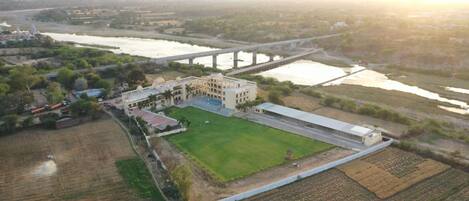 Vista aérea de un edificio grande con un patio central, rodeado de campos y un río. Un puente cruza el río al fondo.