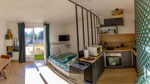 A compact studio apartment with a bed, kitchenette, dining area, and a sliding glass door leading to a balcony.