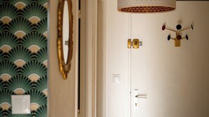 A hallway with a patterned wallpaper, a mirror, a door with a light switch, and a hanging light fixture.