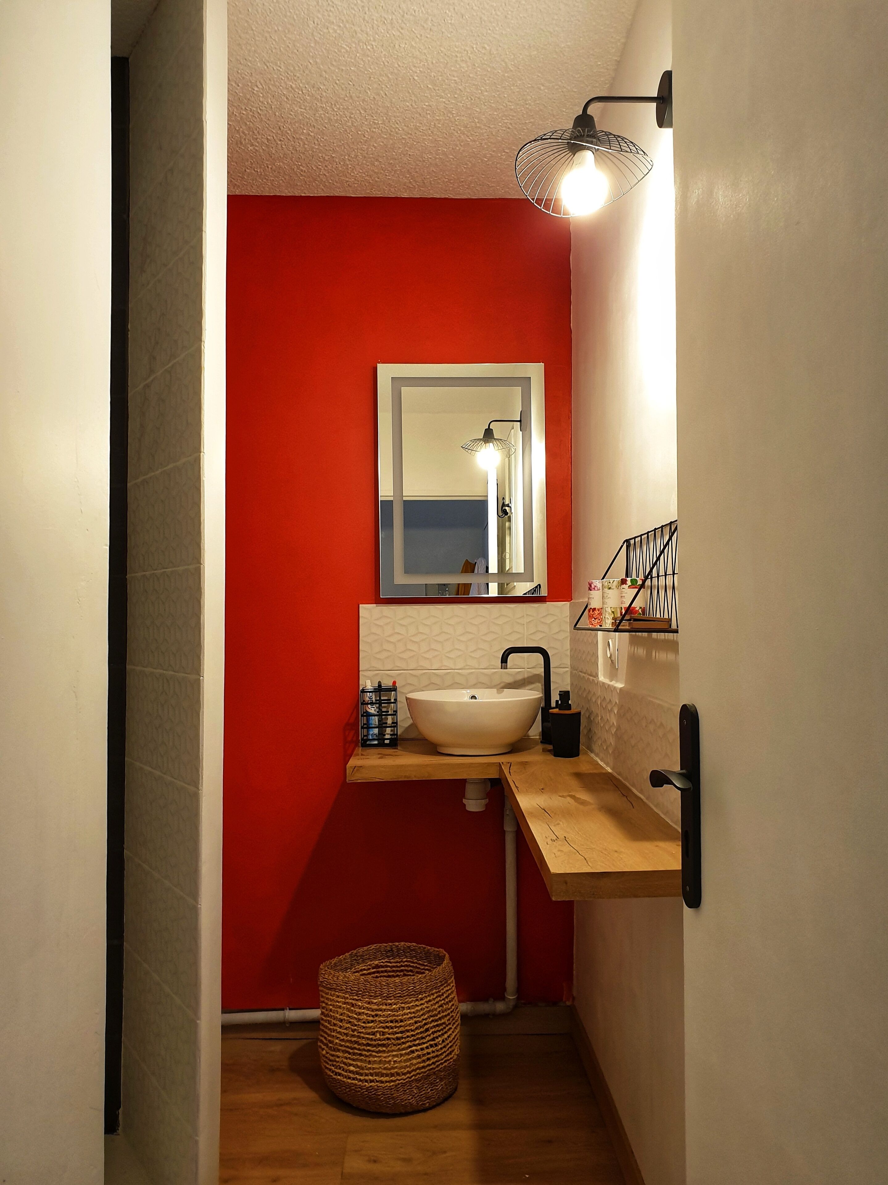 Une petite salle de bain avec un mur d’accent rouge, un lavabo blanc, un miroir et un petit panier.
