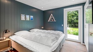 A bedroom with a bed, a nightstand, a lamp, a shelf with decorative items, and a view of greenery through a glass door. - "Baggi" - 900m from the sea by Interhome (Blåvand)