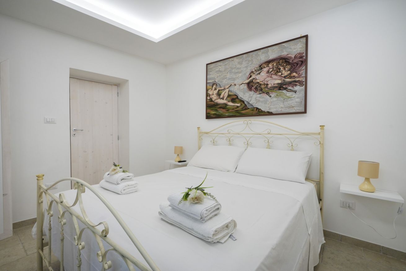 A bedroom with a bed, a painting, a door, and a nightstand with a lamp.