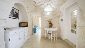 A narrow hallway with white stone walls, a white table with chairs, a vintage radio, and a framed picture on the wall.