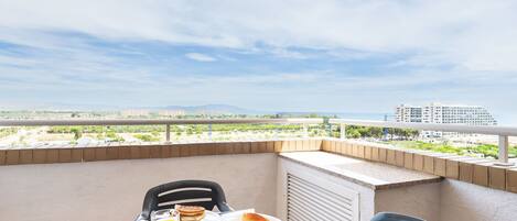 A balcony with breakfast setup including toast, pancakes, and orange juice, overlooking a cityscape with buildings and greenery.