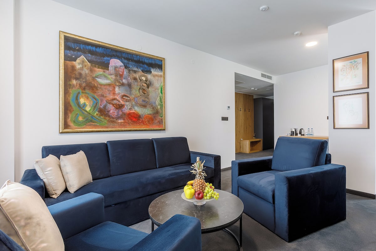 A modern living room with a dark blue sofa, a round coffee table with fruit, and a large abstract painting on the wall.