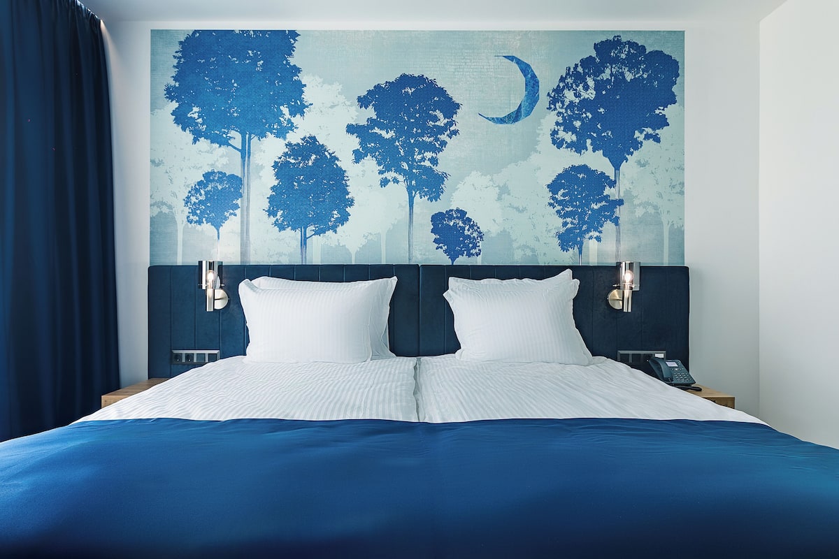 A neatly made bed with white linens and a blue bedspread, flanked by two bedside tables with lamps, in a room with a large wall mural of trees and a crescent moon.
