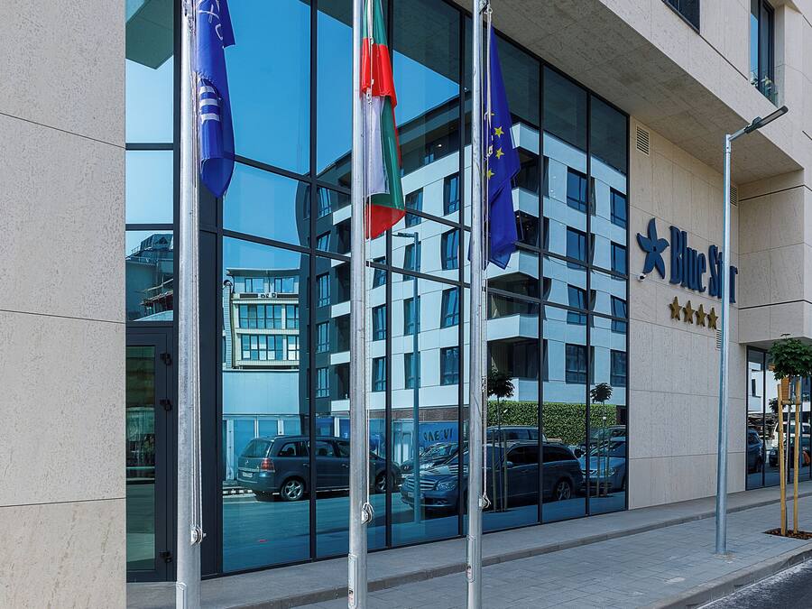 Ein Hotel mit moderner Glasfassade, mehreren Flaggen und freiem Blick auf geparkte Autos vor dem Haus.
