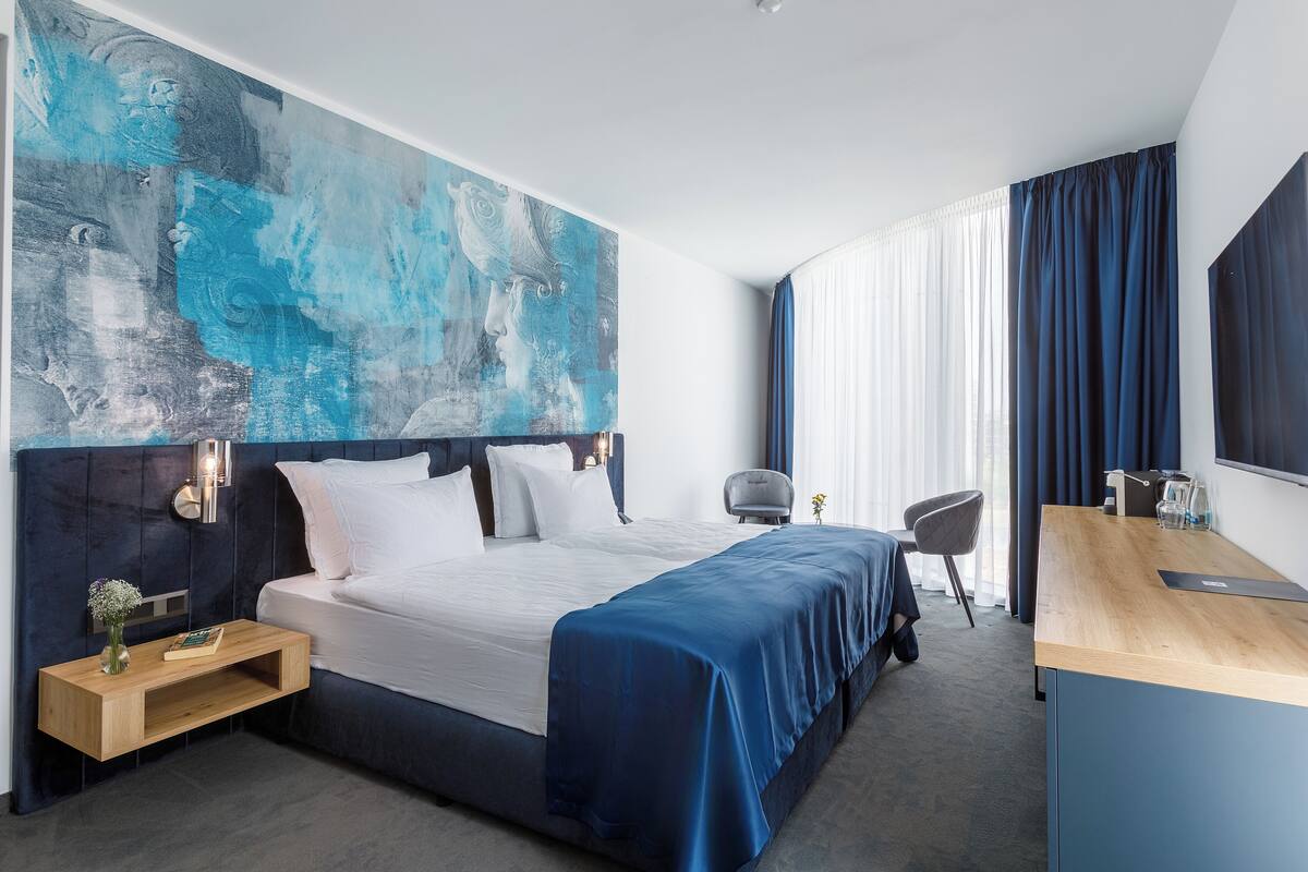 A modern hotel room with a large bed, a wooden desk, a chair, and a large abstract painting on the wall.