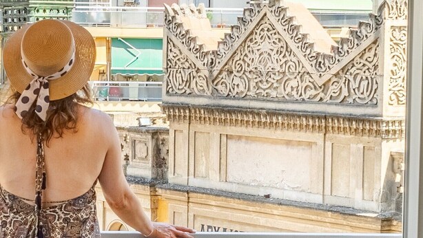 A person standing on a balcony with ornate metal railings, overlooking a courtyard with a stone balustrade and a wooden chair.
