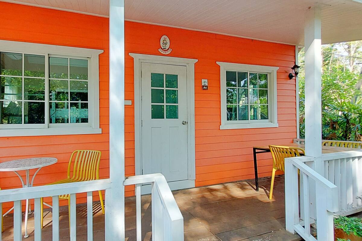 A bright orange building with white trim, a front door, and two windows. There are yellow chairs and a small table on the porch.