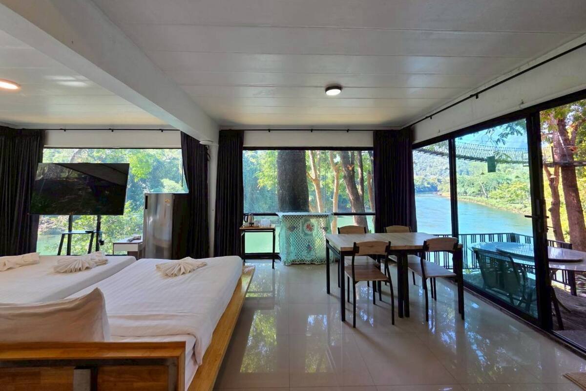 A hotel room with a bed, a TV, a dining area with chairs and a table, and large windows offering a view of a river and trees.