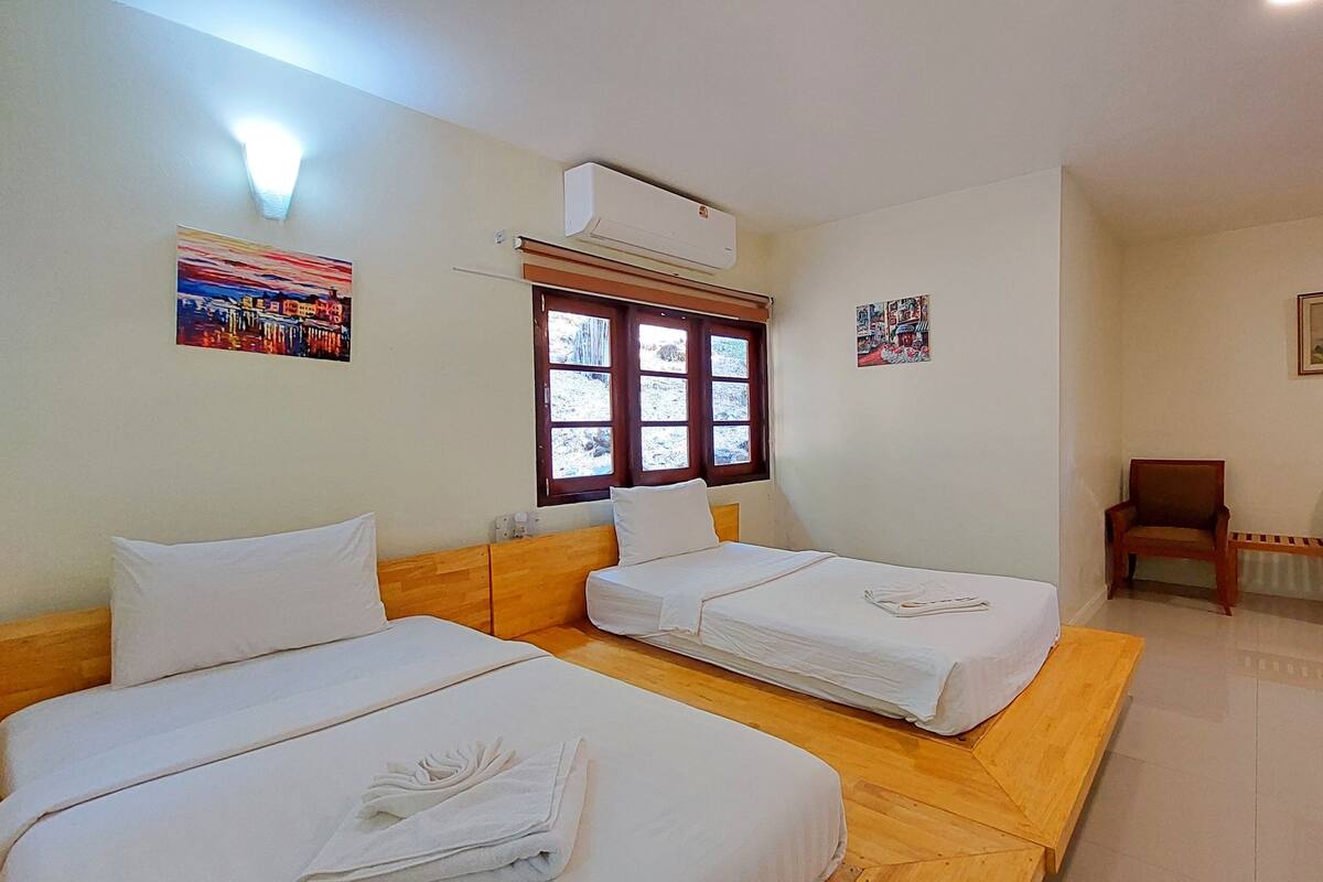 A room with two single beds, a window with a view, a chair, and a wall-mounted air conditioner.