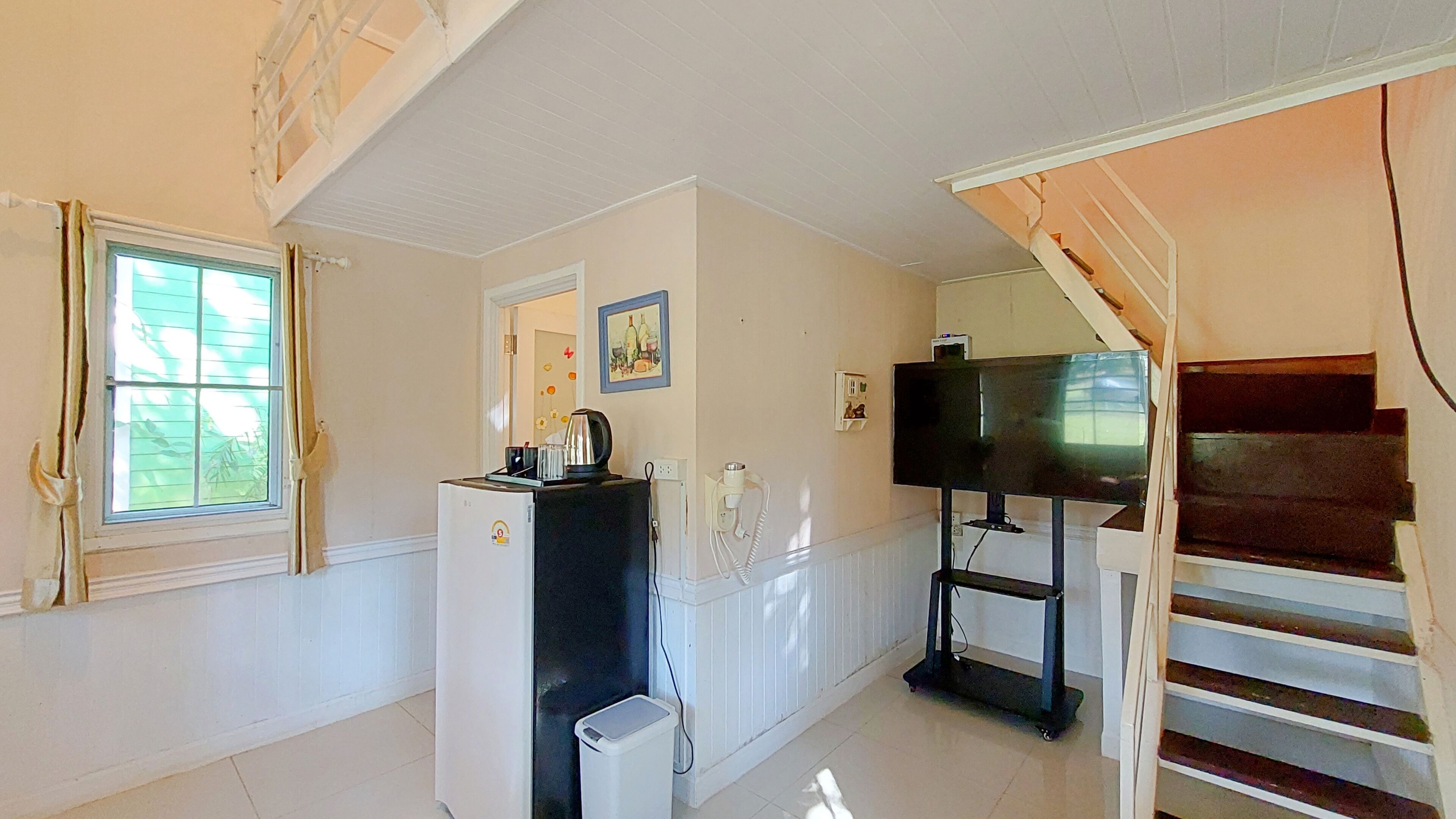 A room with a refrigerator, a television on a stand, a window with curtains, and a staircase.