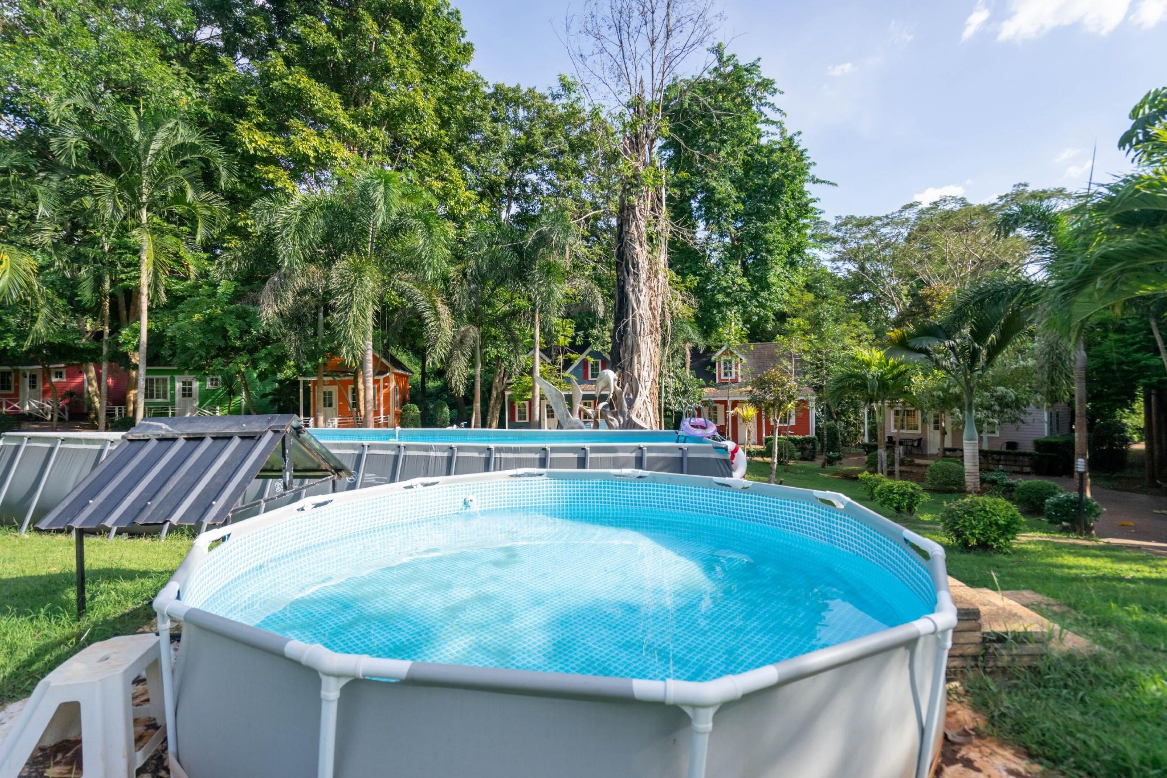 Una piscina rodeada de abundante vegetación y árboles, con una vista de un edificio rojo al fondo.