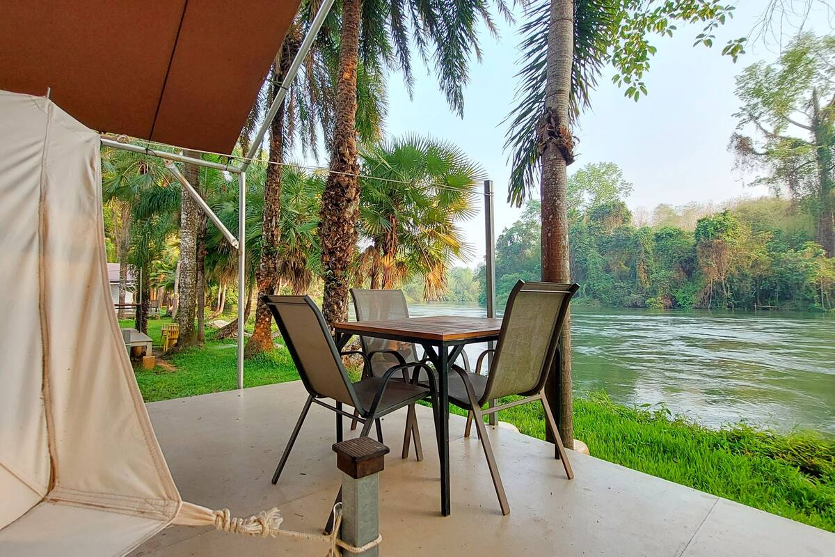 A tented area with a table and chairs overlooking a river, surrounded by palm trees and greenery.