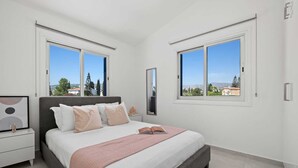 A bedroom with a bed, bedside tables, a mirror, and two windows offering a view of the outside. - Adamante Luxury Villa by Nomads (Paphos)
