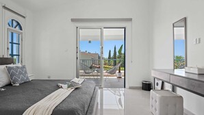A bedroom with a bed, a bedside table, a dresser, a mirror, and a balcony with chairs and a potted plant. - Adamante Luxury Villa by Nomads (Paphos)