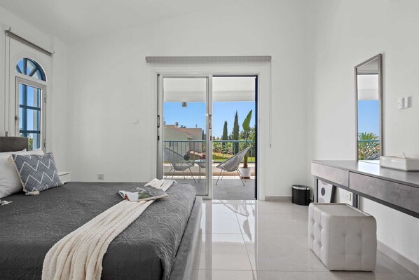 A bedroom with a bed, a bedside table, a dresser, a mirror, and a balcony with chairs and a potted plant.