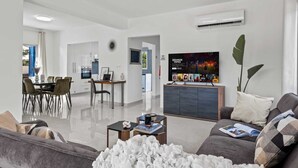 A modern living room with a sofa, coffee table, and a TV mounted on the wall. There is a dining area with a table and chairs, and a kitchen visible in the background. - Adamante Luxury Villa by Nomads (Paphos)