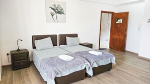 Egy franciaágyas hálószoba, rajzolt mintájú ágytakaróval, két éjjeliszekrény, egy kisebb asztal, egy falra szerelt lámpa és egy ajtó, amelyen a „Legyen otthonunk, legyen jó otthonunk” felirat látható.