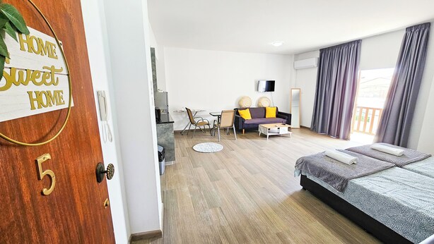 Egy modern hotelszoba, melyben fa padló, mintás ágytakaróval ellátott ágy, párnákkal teli kanapé, egy kis asztal és egy szék található. Az ajtón egy tábla van, amelyen a következő felirat olvasható: „OTTHON, édes OTTHON”.