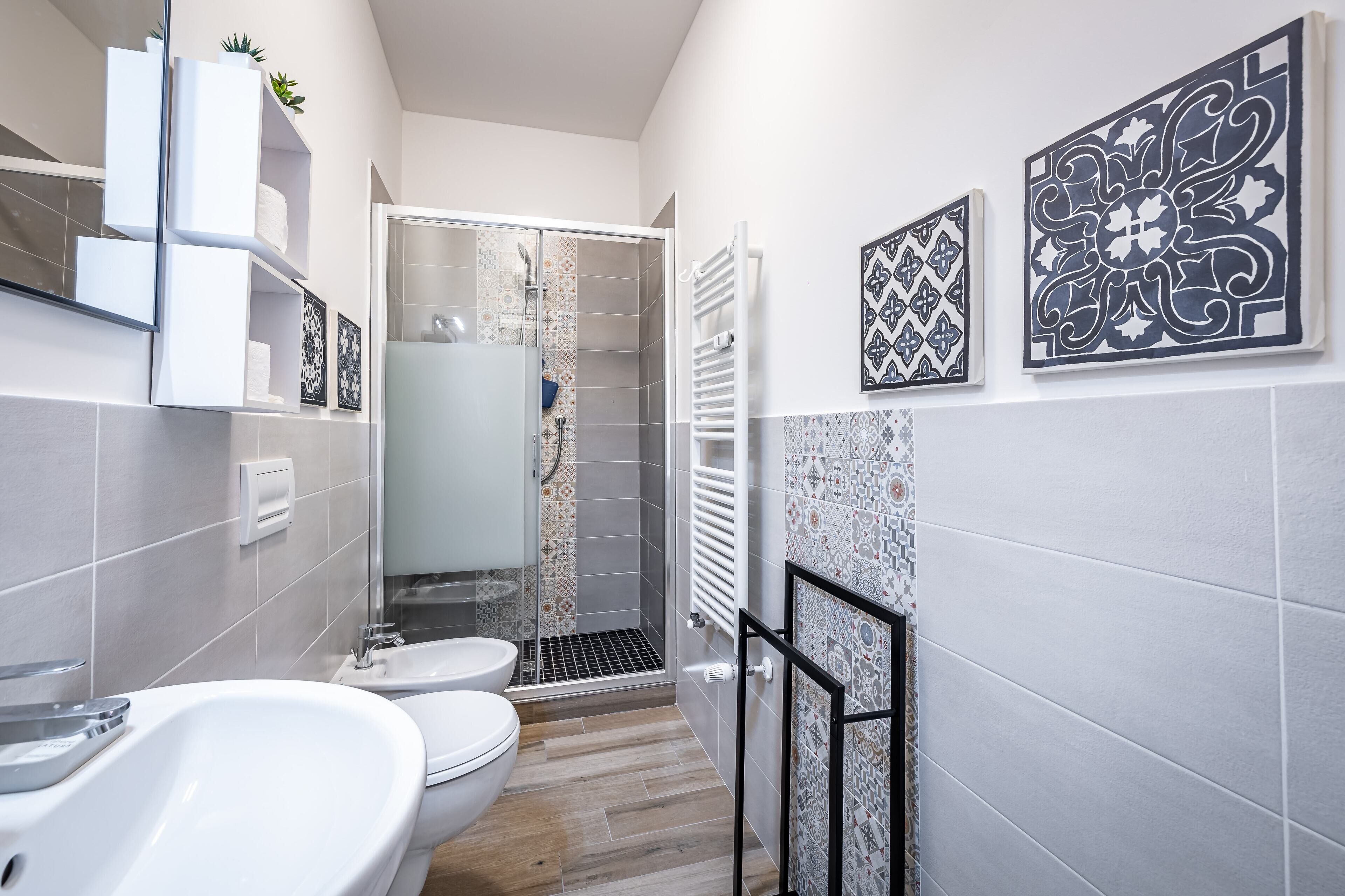 A modern bathroom with a white sink, toilet, and shower area. There are decorative tiles on the walls and a glass shower enclosure.