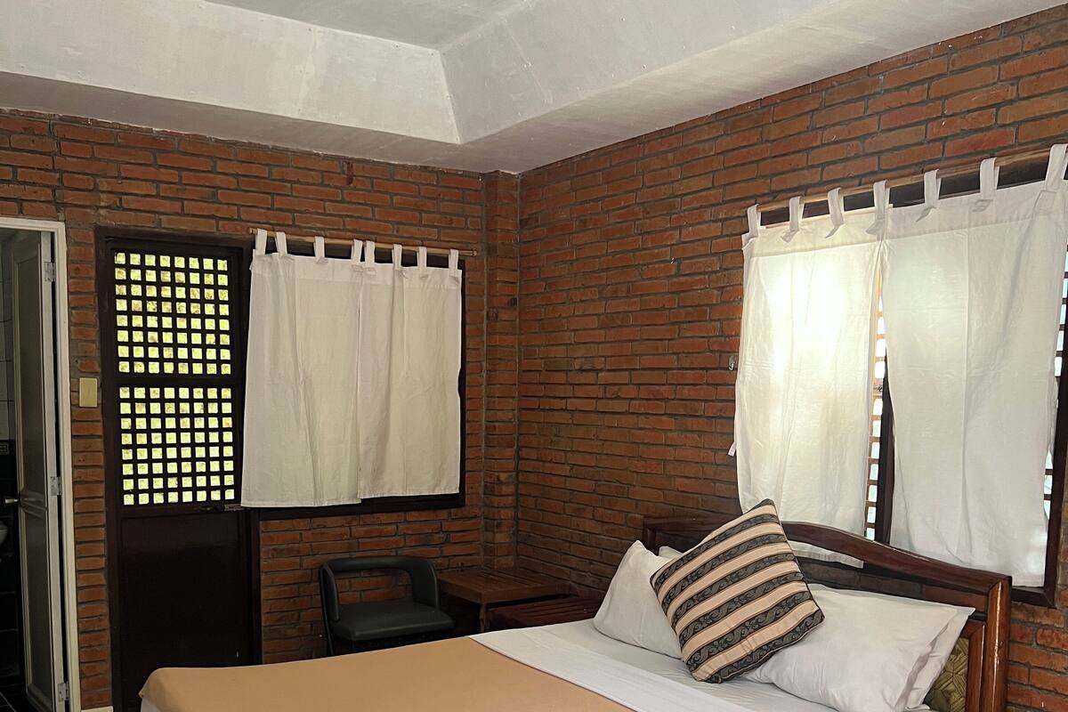 A room with a bed, a chair, a table, and a brick wall.