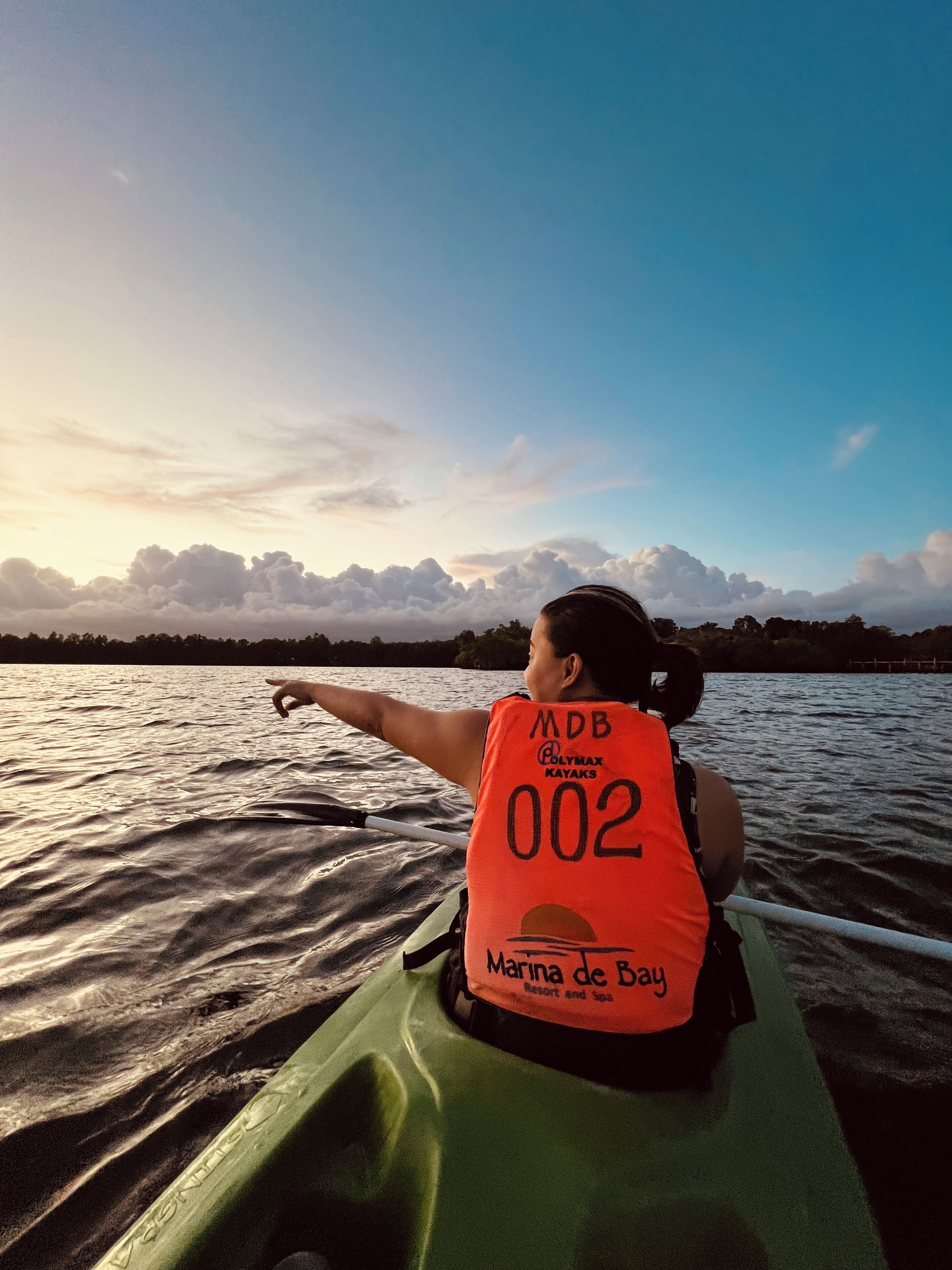 Una persona en kayak, con chaleco salvavidas con el número 002, señalando hacia el agua.