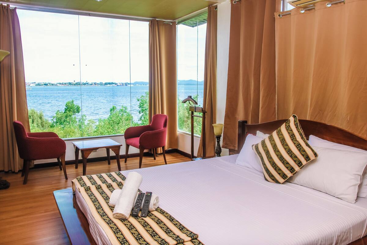 A hotel room with a large bed, two red chairs, a small table, and a view of a body of water.